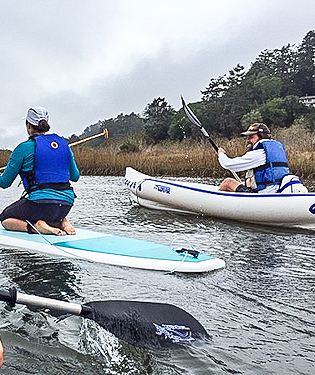 Two people paddling in a calm marsh area: one on a stand-up paddleboard and the other in a kayak.
