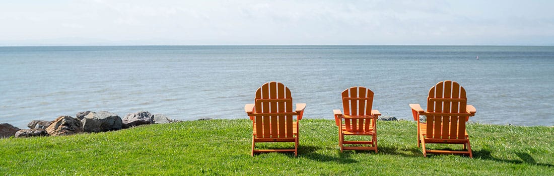 Three empty but inviting Adirondack chairs sit on a green lawn overlooking the bay.